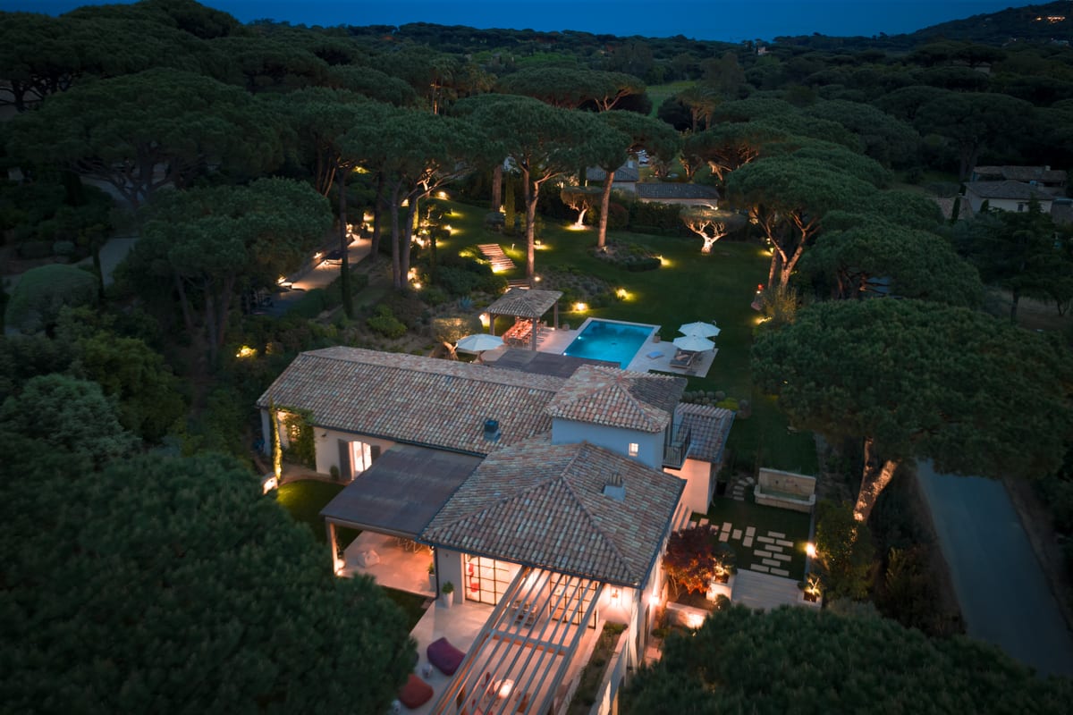 The garden from the air at dusk, lit path by path, the pool glowing between the pines.