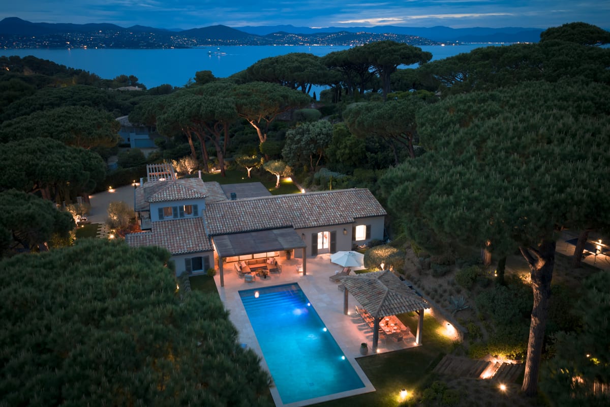 The villa seen from the air at dusk, lit among umbrella pines, with the pool below and the bay beyond.