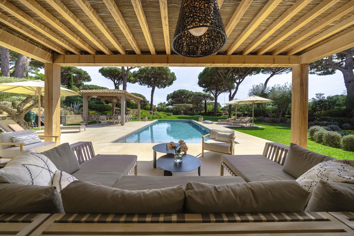 Deep sofas under the timber pergola, woven pendant lamps overhead, the pool beyond.