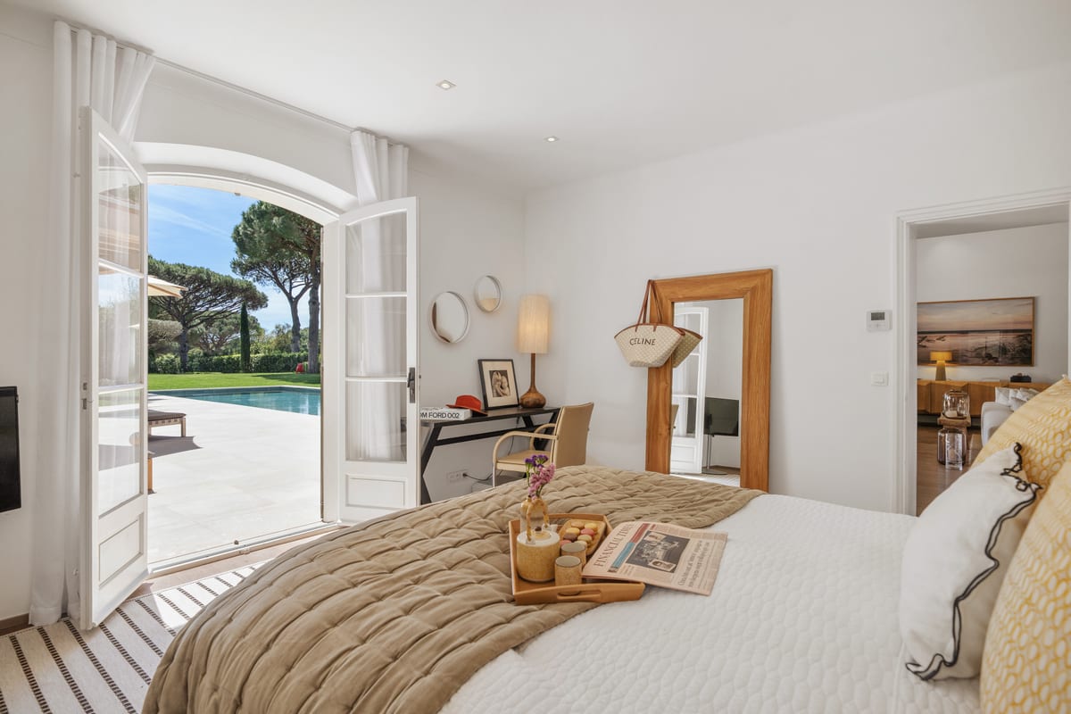 A bedroom with double doors open onto the garden, a striped rug and a made bed.