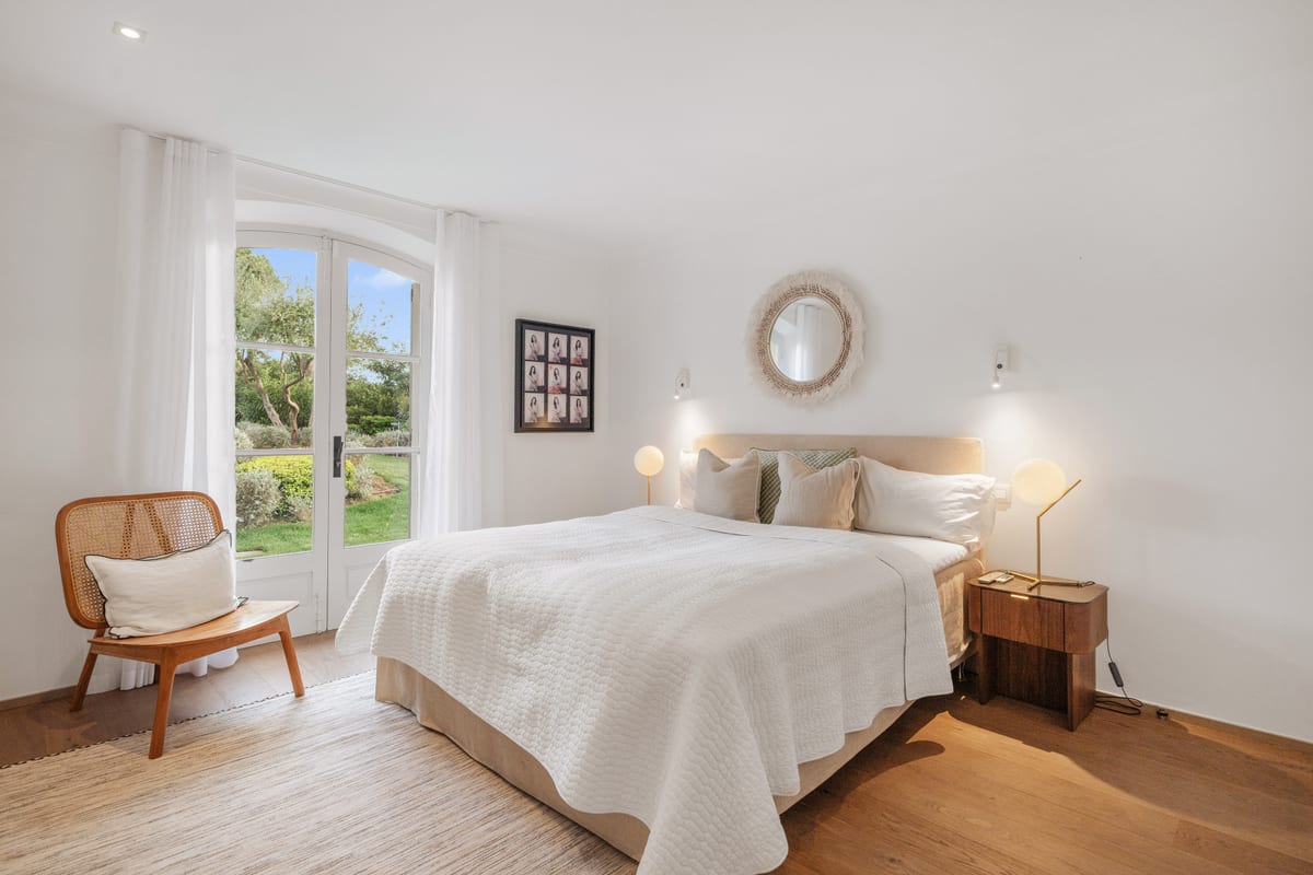 A white bedroom with a cane armchair, a round mirror and French doors.