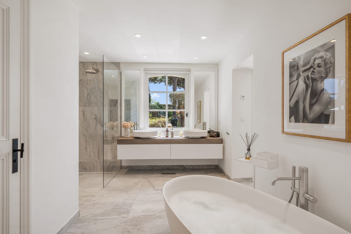 A bathroom in pale marble with a freestanding tub under a window looking onto greenery.