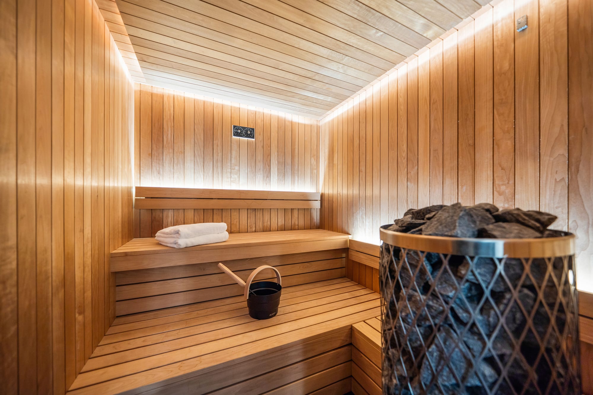 A timber-panelled sauna with a stove, benches and indirect lighting along the ceiling.