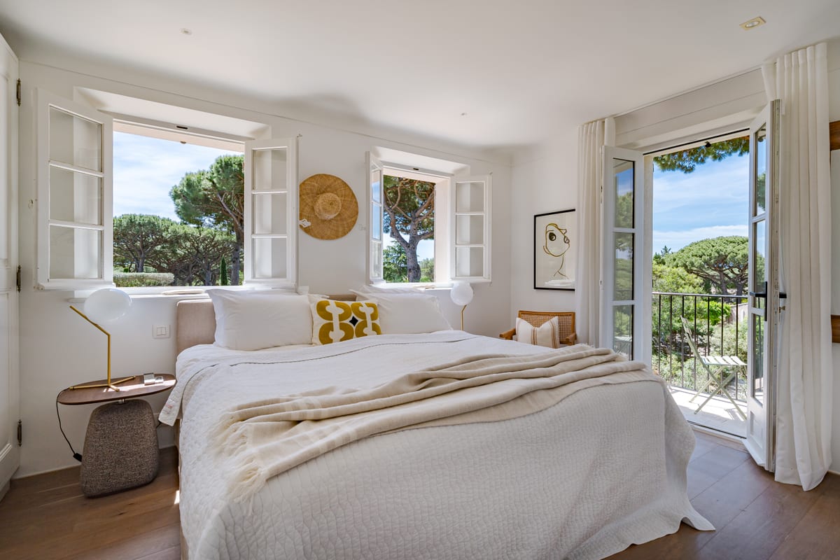 A bedroom with shuttered windows, a pine visible outside and a door open to the garden.
