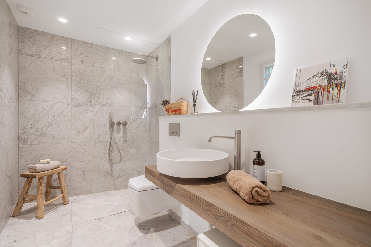 A marble bathroom with a walk-in shower, a round mirror and a basin set on timber.