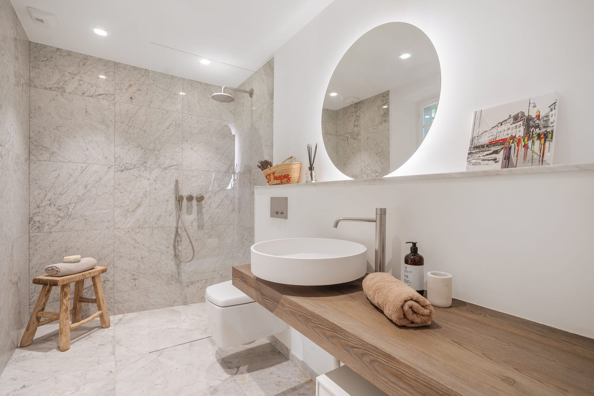 A marble bathroom with a walk-in shower, a round mirror and a basin set on timber.