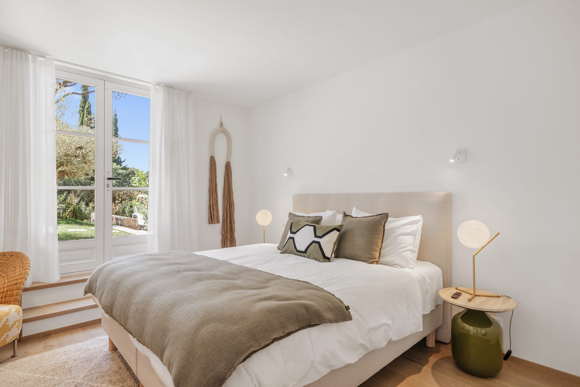 A bedroom in pale linen, a low chair by the window and umbrella pines outside.