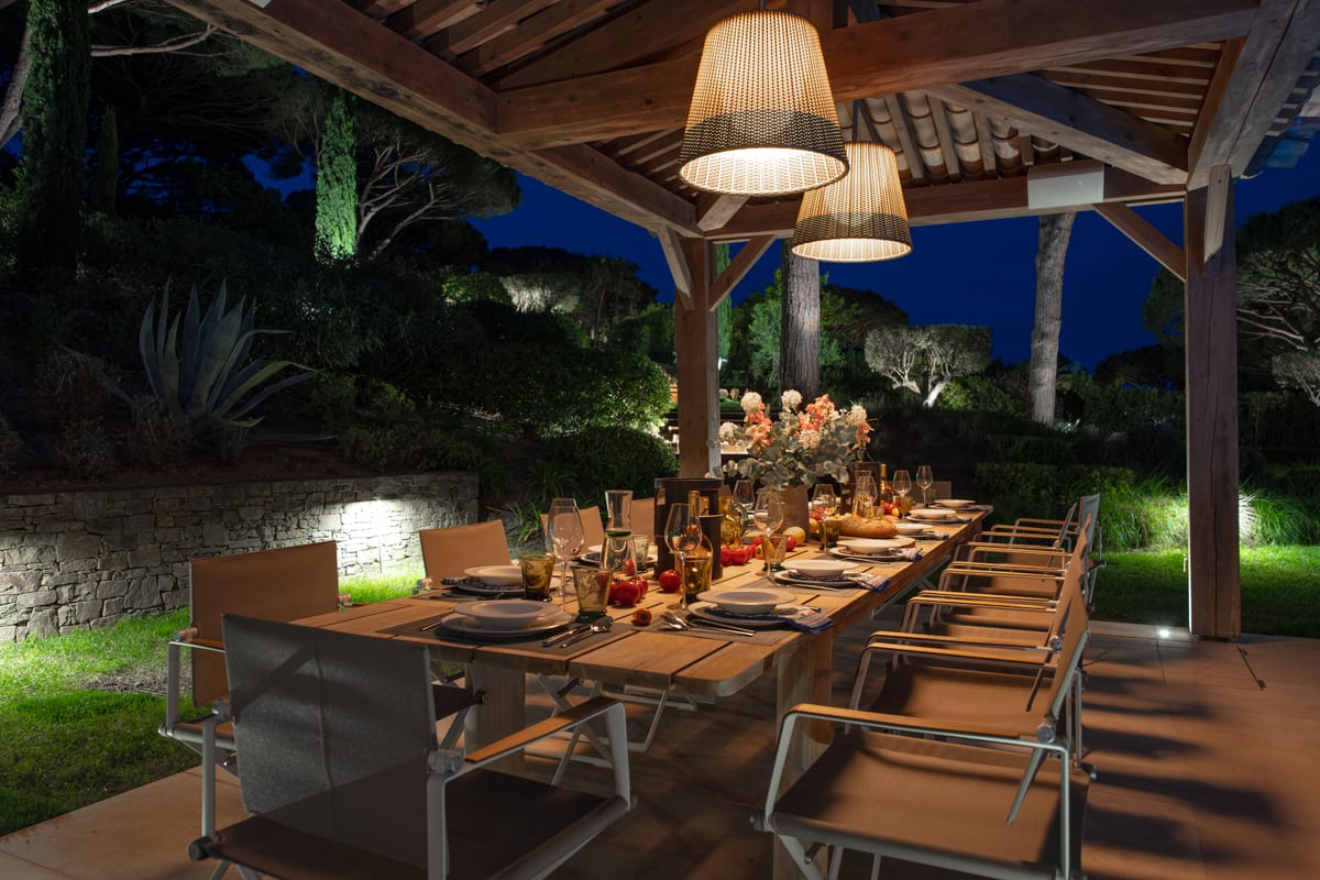 A long outdoor table laid for dinner under the pergola, lit from above, the garden dark behind.