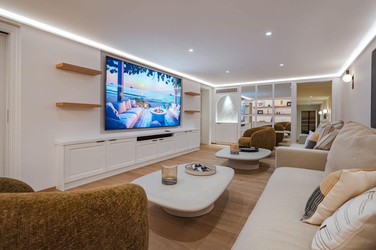 A television room: a wide wall-mounted screen above low white cabinets, two curved coffee tables, deep sofas and armchairs, with cove lighting along the ceiling.