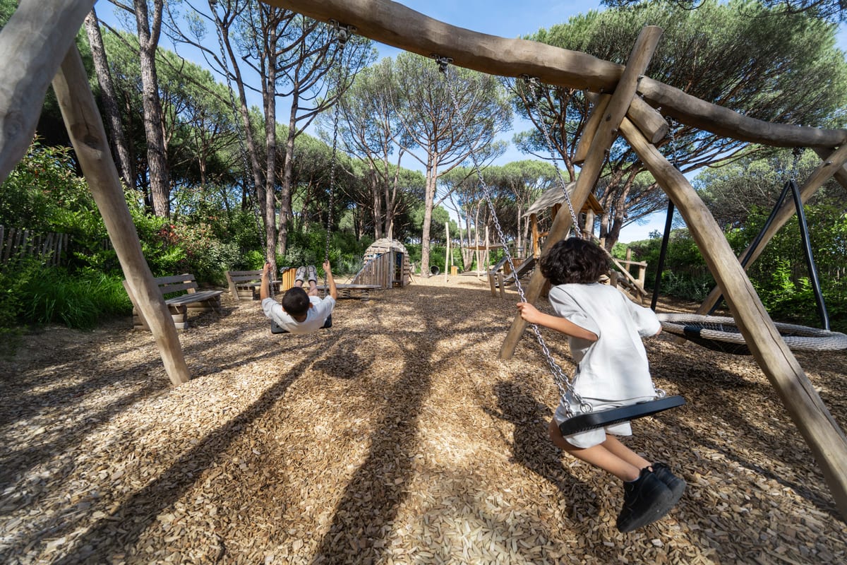 A rope-and-timber swing frame under the umbrella pines, a child running through the dappled shade.