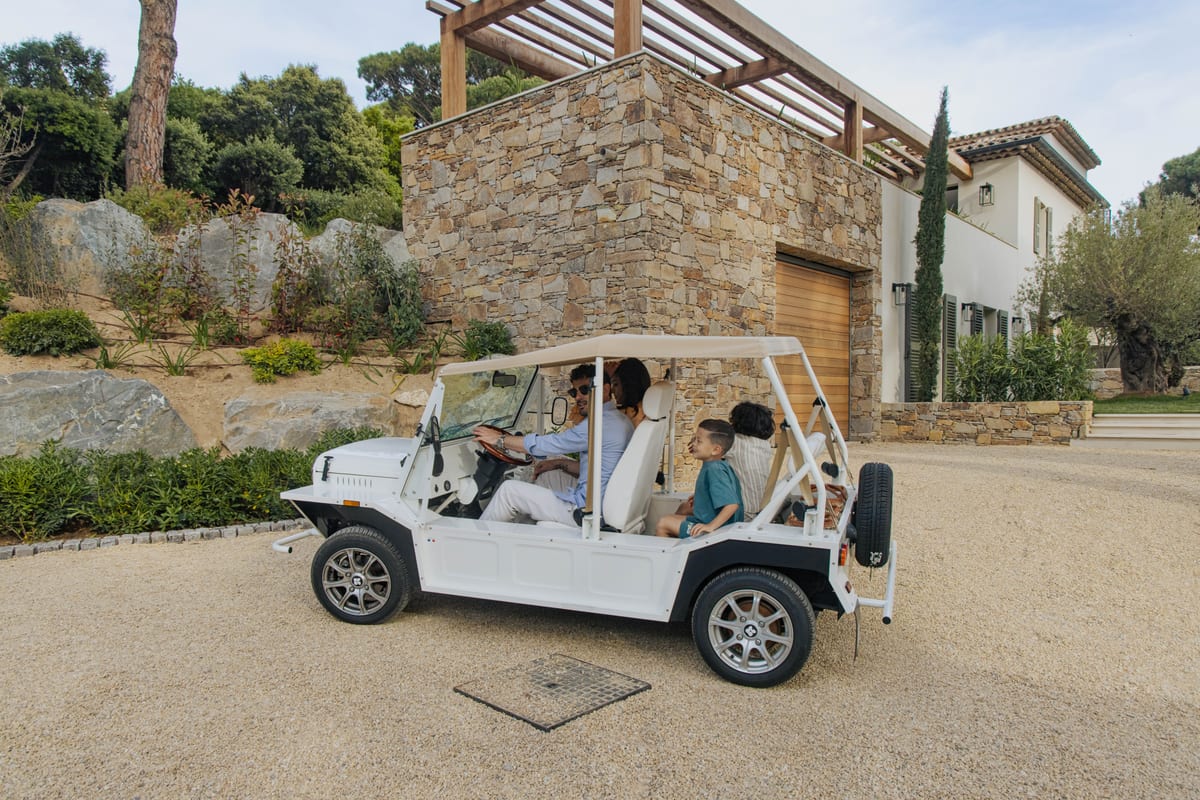A white open-sided Moke parked on the gravel drive in front of a dry-stone wall, with people aboard.