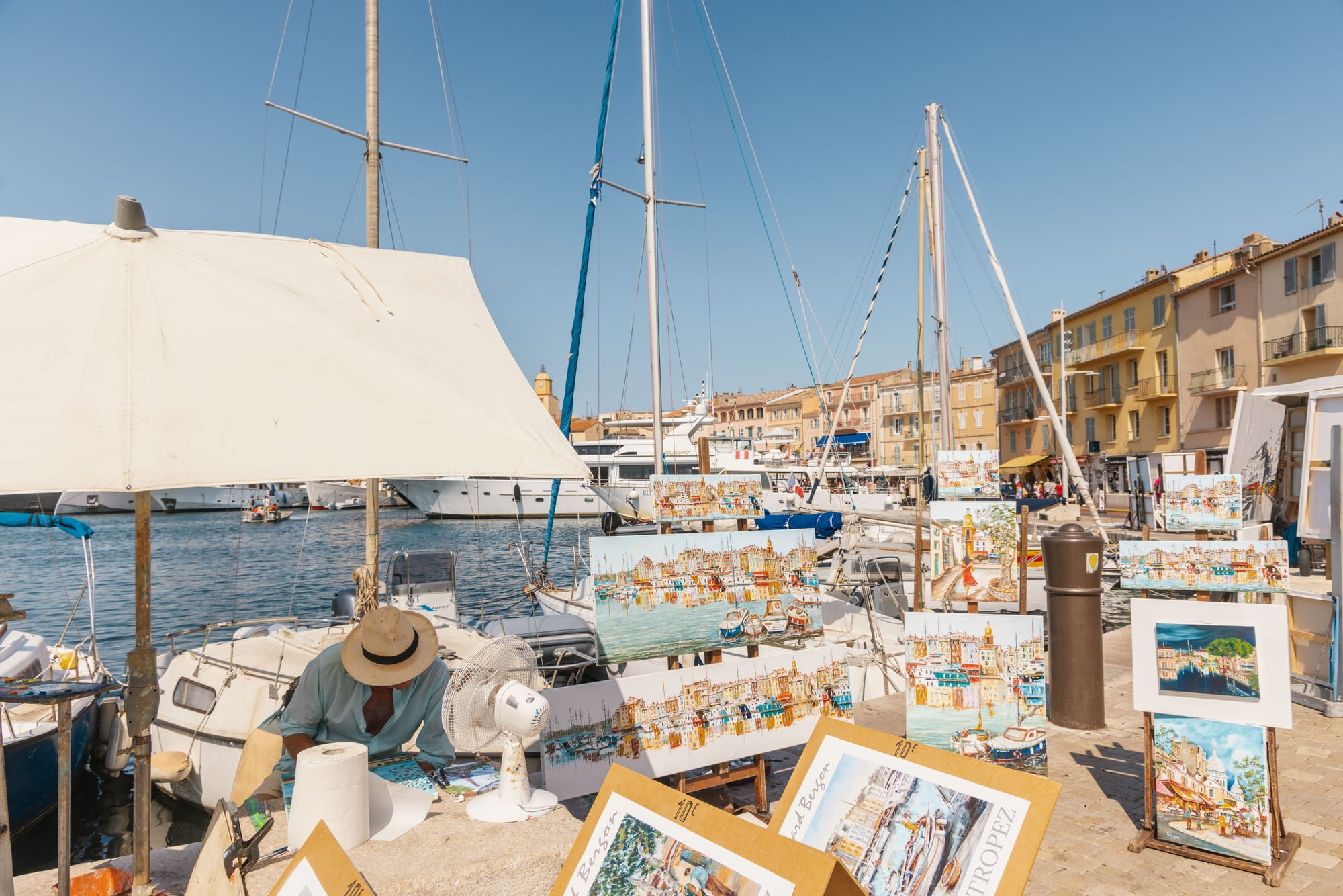A painter's stall on the quay, canvases propped along the ground, a boat and a parasol behind.