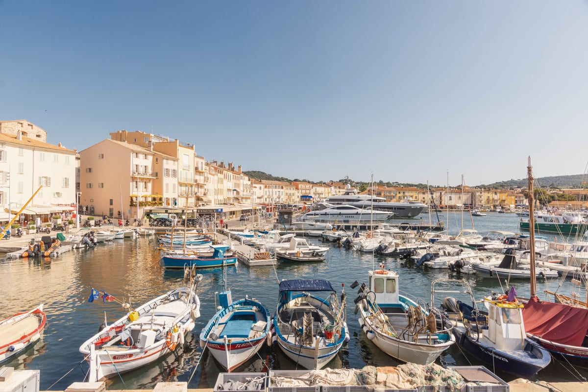 The old port of Saint-Tropez: painted wooden fishing boats moored in the foreground, ochre facades and the bell tower behind, large yachts along the far quay.